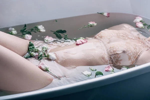 Cropped view of sexy woman in beige lace underwear lying in clear water with flowers in white bathtub — Stock Photo