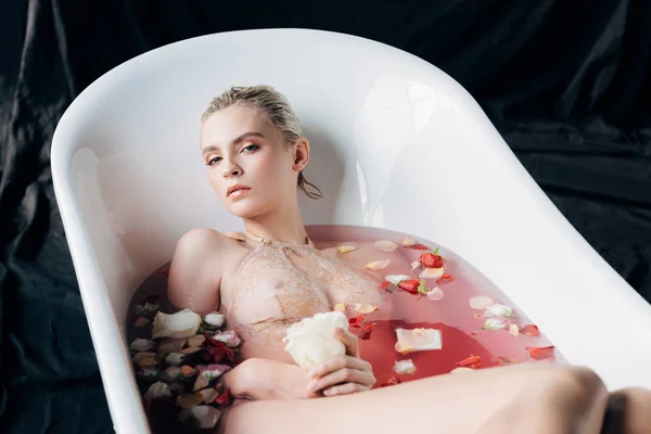 Hermosa mujer rubia húmeda y sexy tumbada en la bañera con agua rosa y pétalos - foto de stock