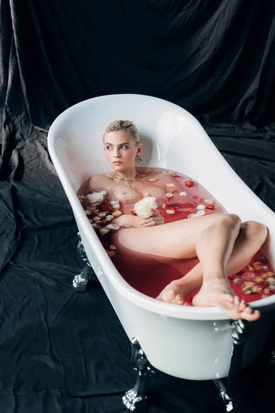 Beautiful barefoot wet and sexy blonde woman lying in bathtub with pink water and petals on black background — Stock Photo