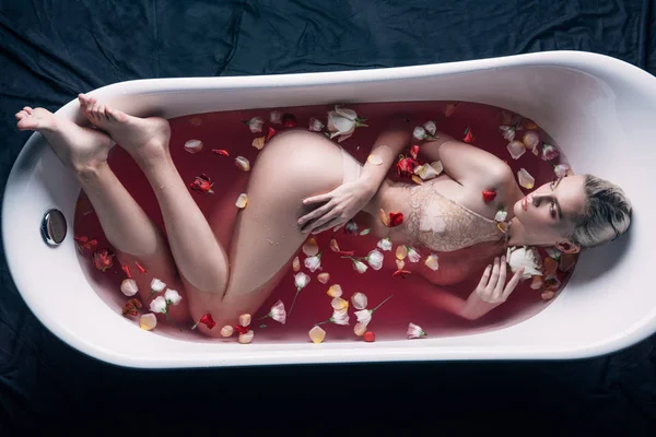 Top view of sexy woman in beige lingerie in bathtub with pink water and petals looking away — Stock Photo