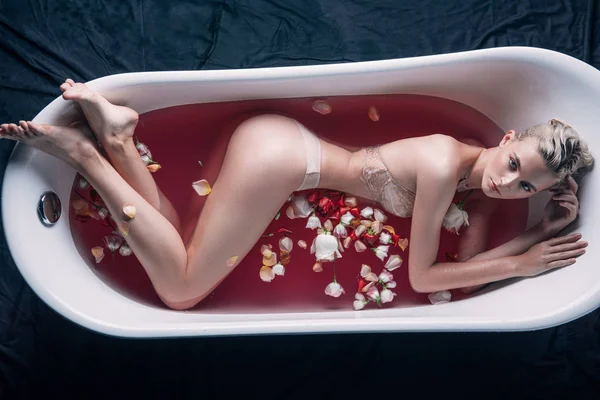 Vista superior de mujer sexy en lencería beige posando en bañera con agua rosa y pétalos - foto de stock