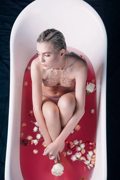 Sexy woman in beige lingerie sitting in bathtub with pink water and rose petals — Stock Photo