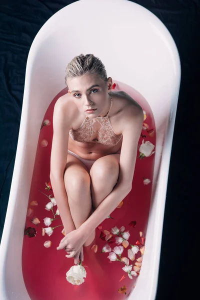Femme sexy en lingerie beige assise dans la baignoire avec de l'eau rose et des pétales — Photo de stock